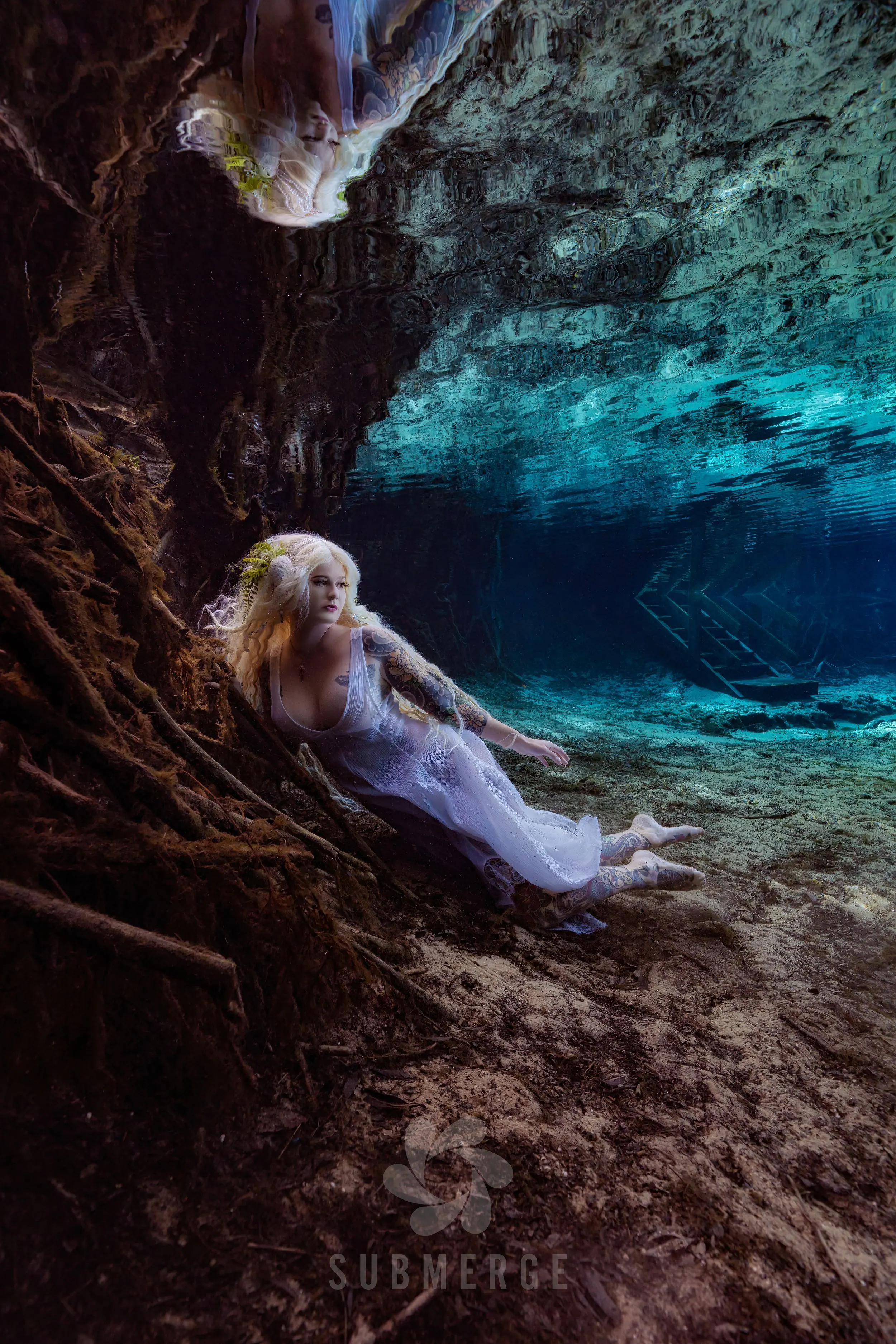 Model in a flowing white gown resting among sunken tree roots in a crystal-clear spring